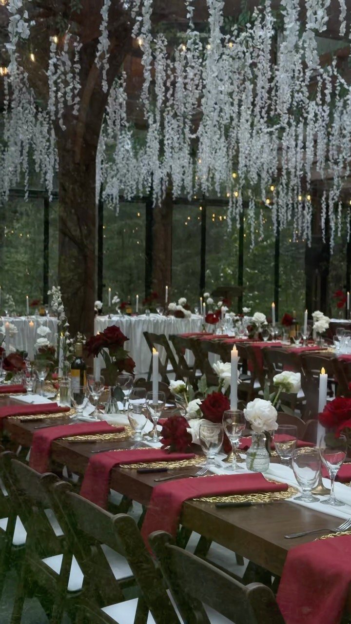 Twilight Forest Wedding Ideas: 10 Concepts for 2026 2 Long wooden dining tables set with red linens and flowers under hanging white floral decorations.