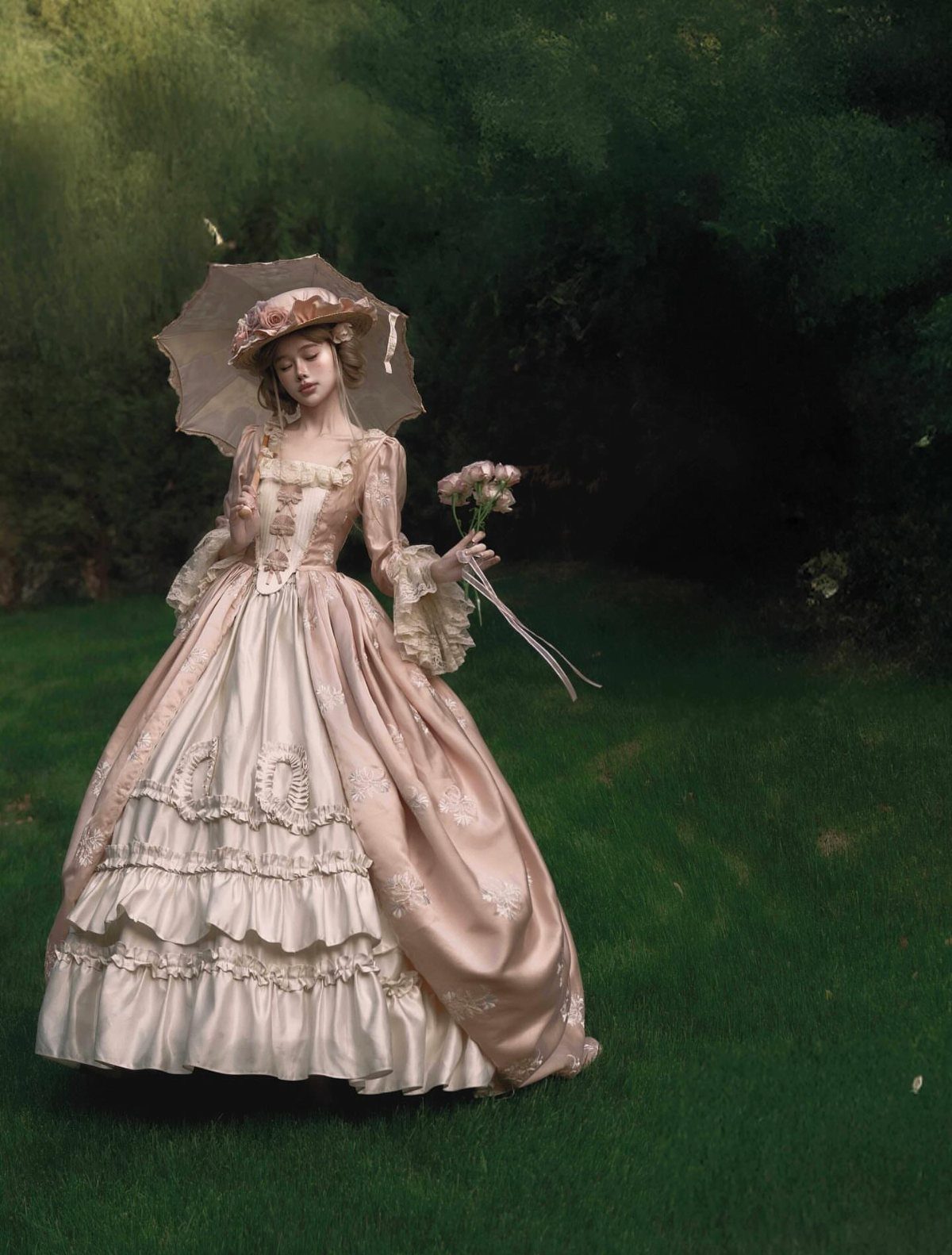 A woman in a pink Victorian gown and hat stands in a green field holding flowers.