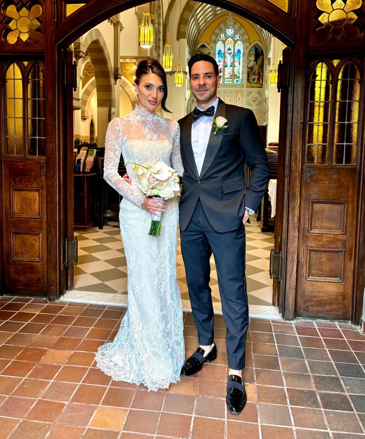 Bride in a long-sleeved lace wedding gown and groom in a tuxedo standing in a church doorway.