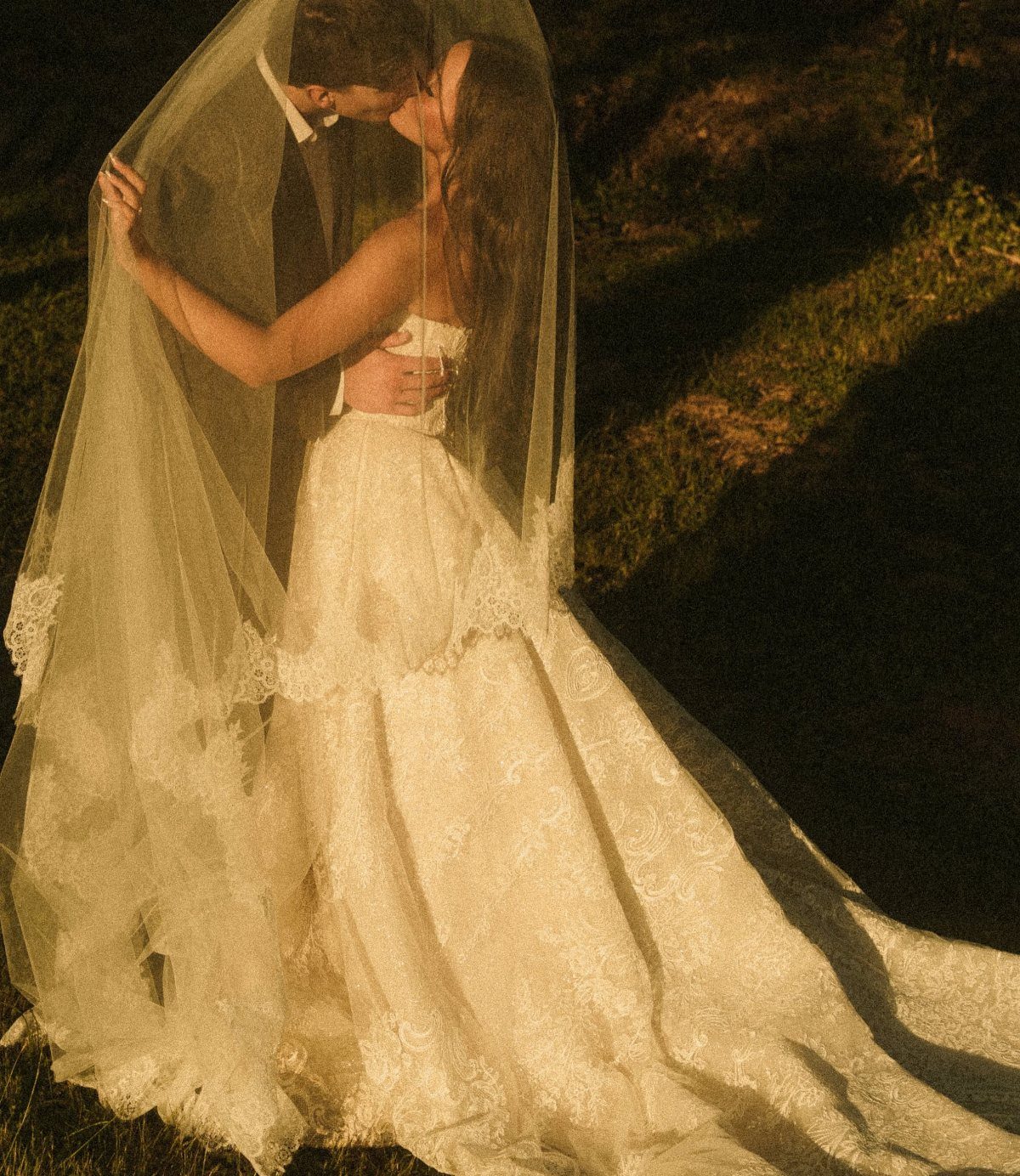 A bride wearing a white lace wedding dress and a long, sheer lace-trimmed veil outdoors.