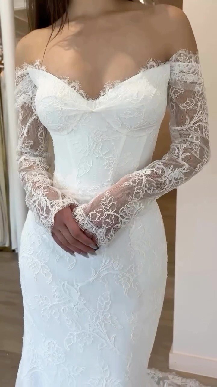 A woman wearing a white lace off-the-shoulder wedding dress with long sheer lace sleeves.