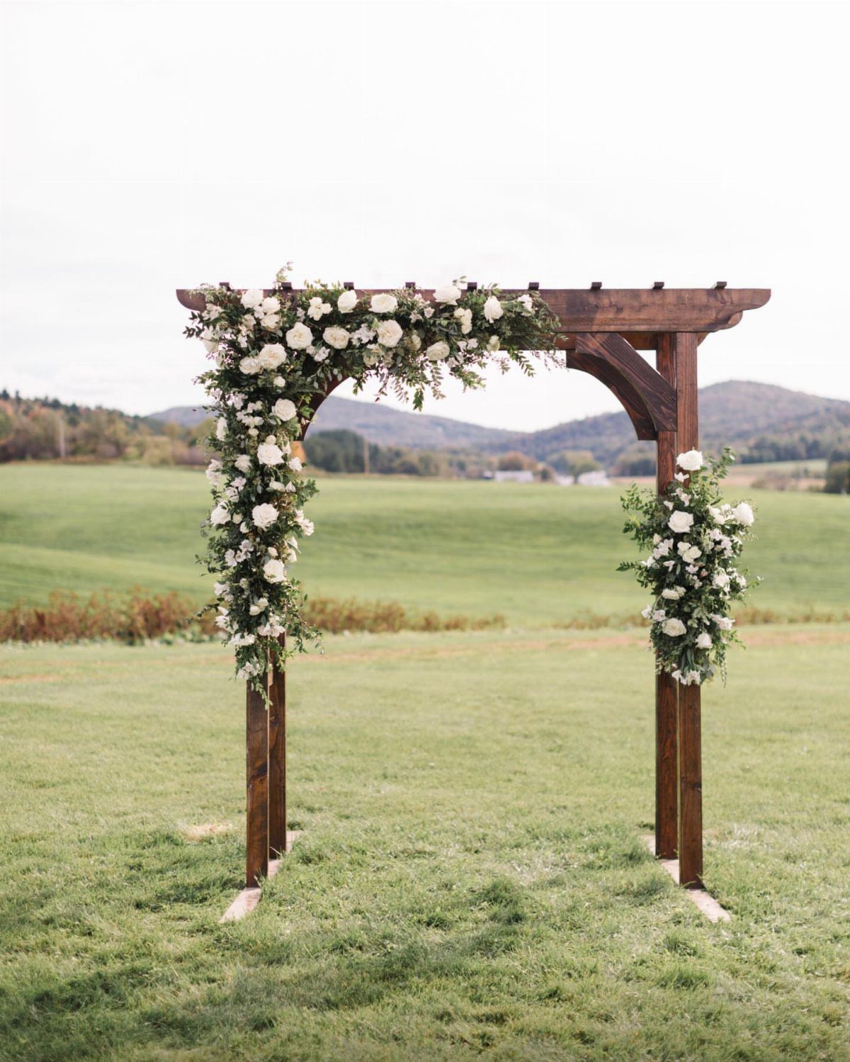 A wooden wedding arbor with white floral arrangements standing in a large green field.