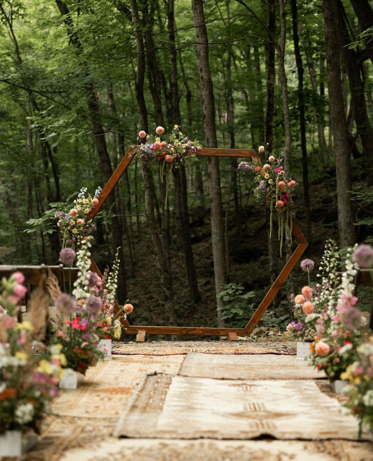 A wooden hexagonal wedding arbor decorated with colorful flowers in a forest setting.