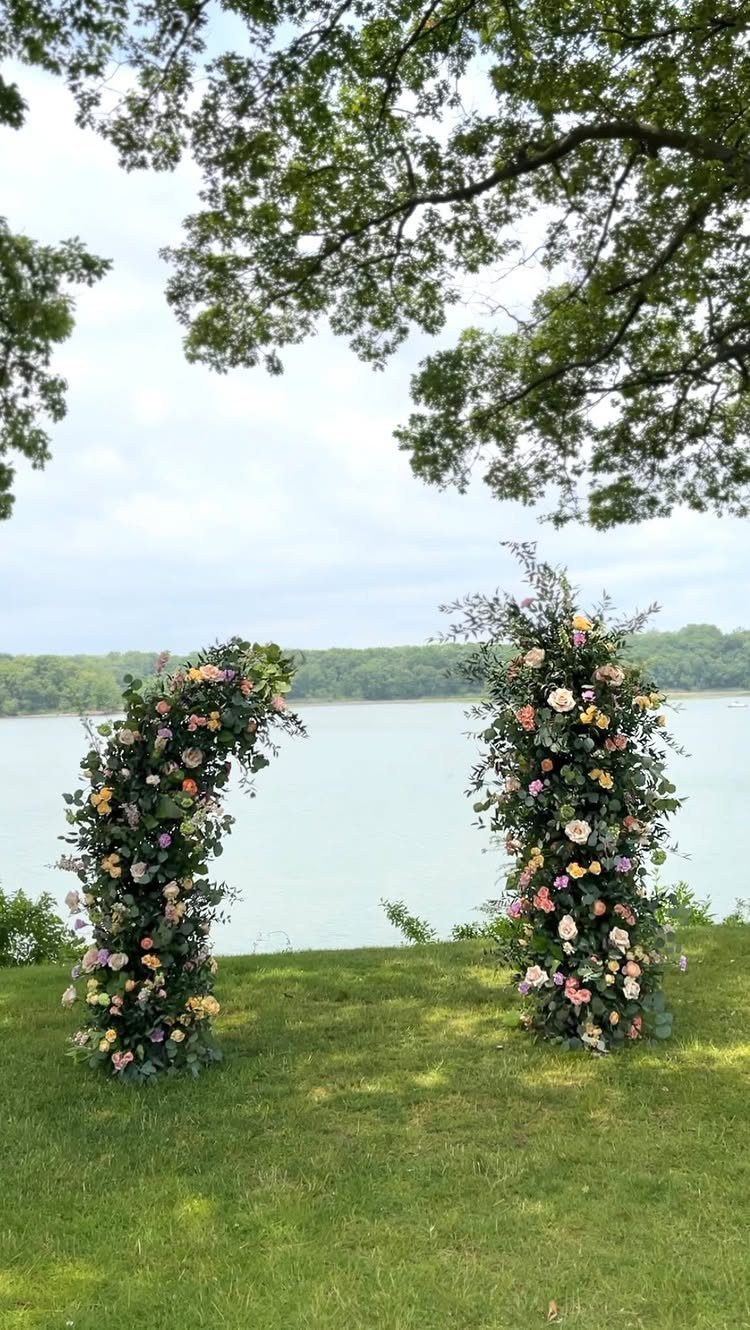 Two tall floral pillars with colorful flowers and greenery standing on a lawn by a lake.