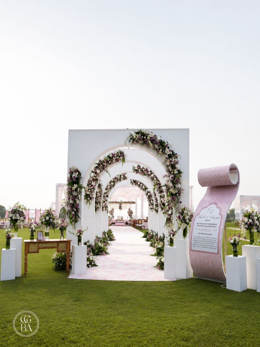 White floral arches create a decorated walkway on a green lawn under a bright sky.