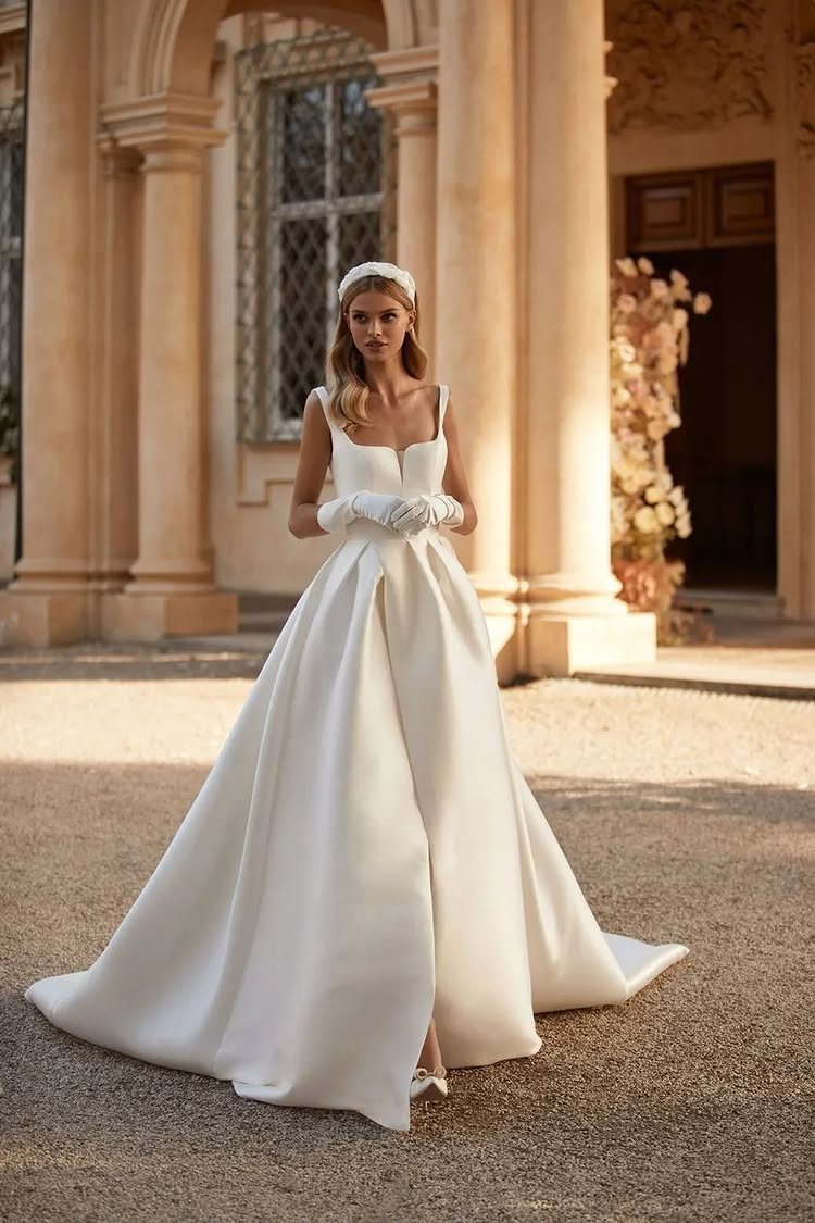 Wedding Dress Trends: 10 Looks for 2026 6 A woman in a minimalist white satin wedding dress standing before stone columns.