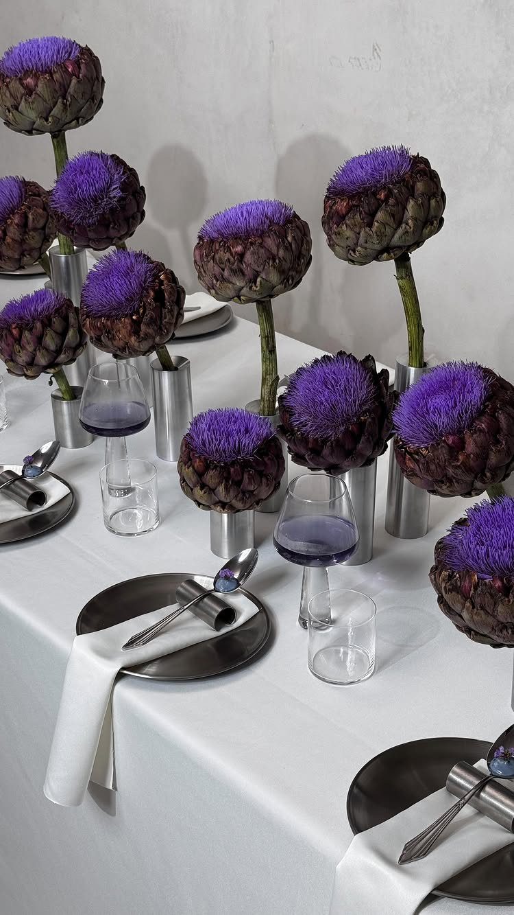 Purple artichoke centerpieces arranged on a white tablecloth with dark plates and wine glasses.