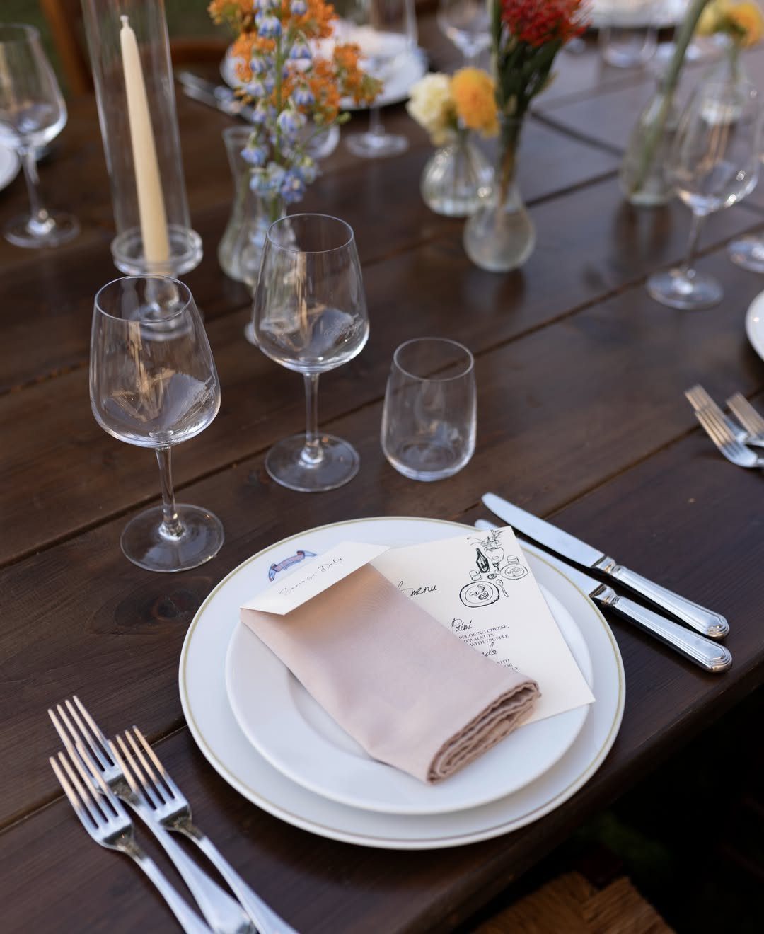 A rustic wooden table set with white plates, pink napkins, silverware, and small wildflower arrangements.