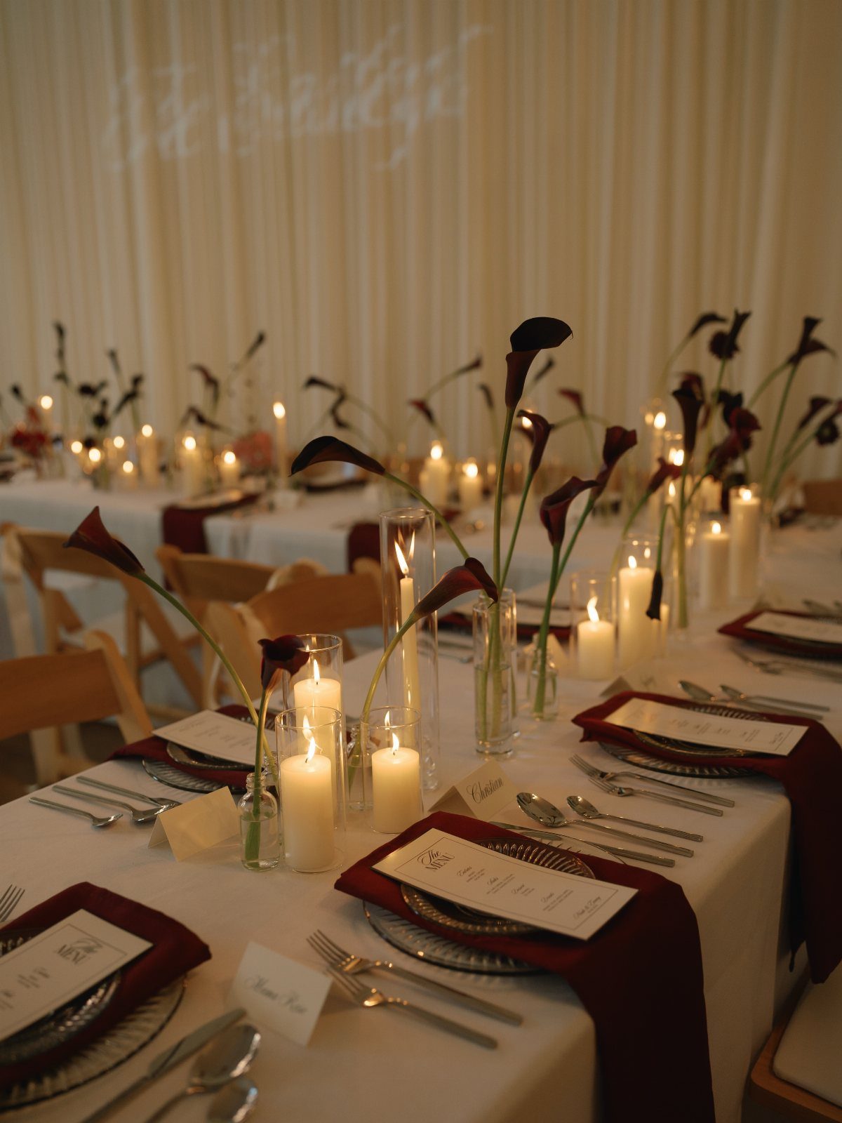 A long wedding reception table decorated with white candles, burgundy napkins, and dark calla lilies.