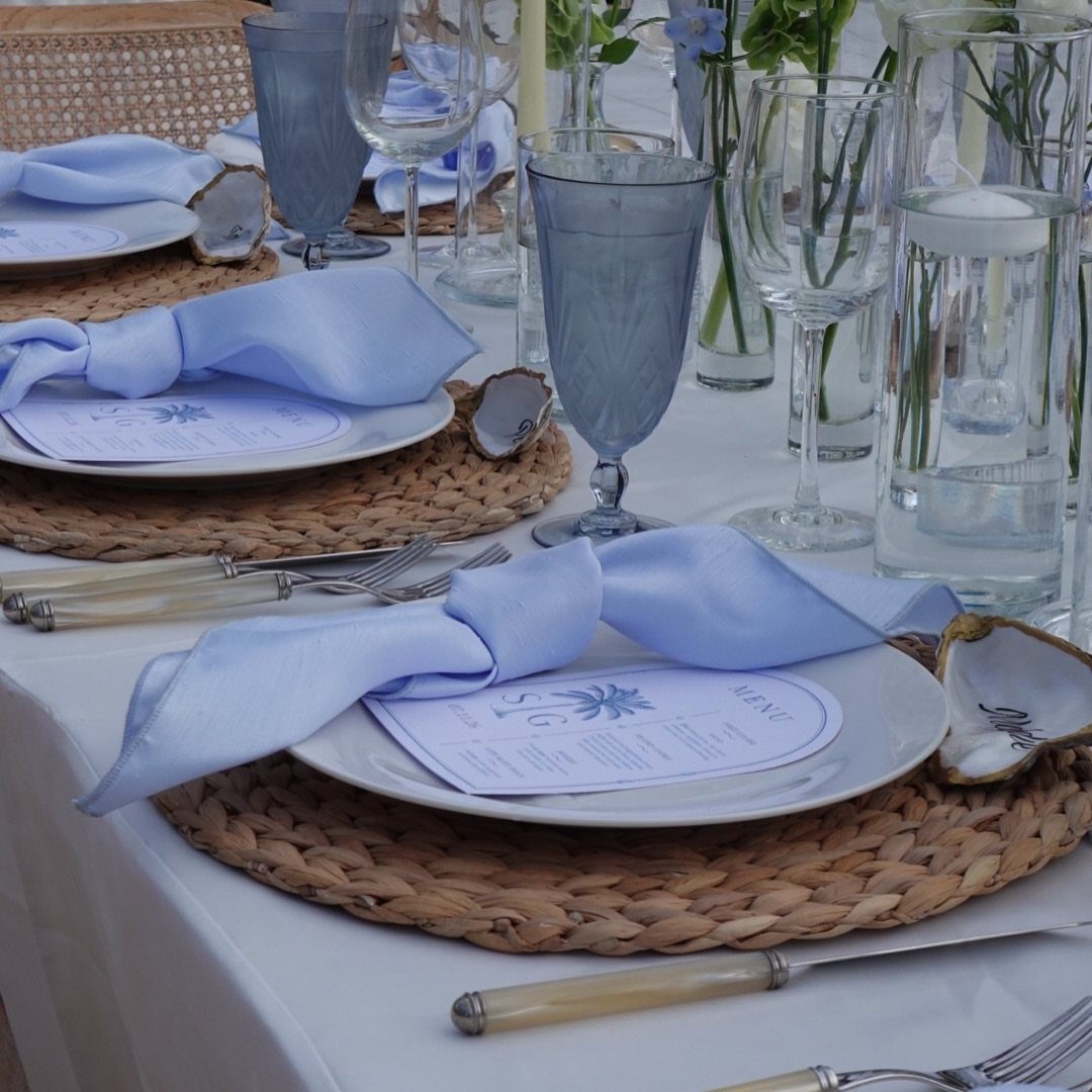 A formal table setting with blue napkins, white plates, and woven placemats on a white tablecloth.