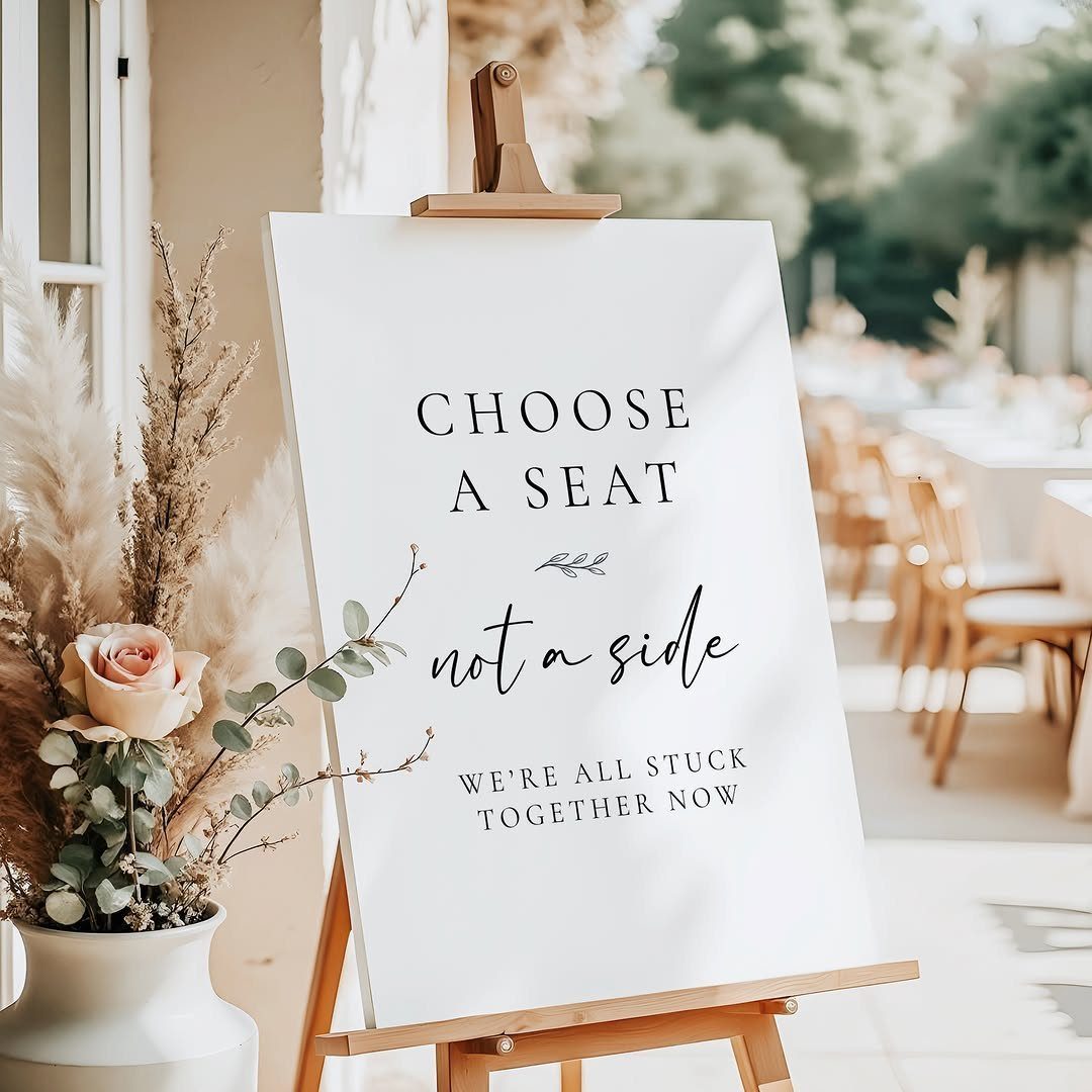 A white wedding sign on a wooden easel with humorous text and dried floral arrangements.