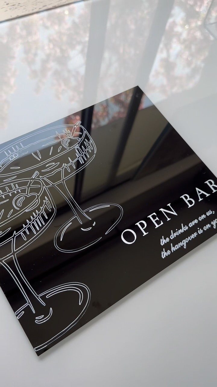 Black acrylic open bar sign with white cocktail illustration and humorous text on a white surface.