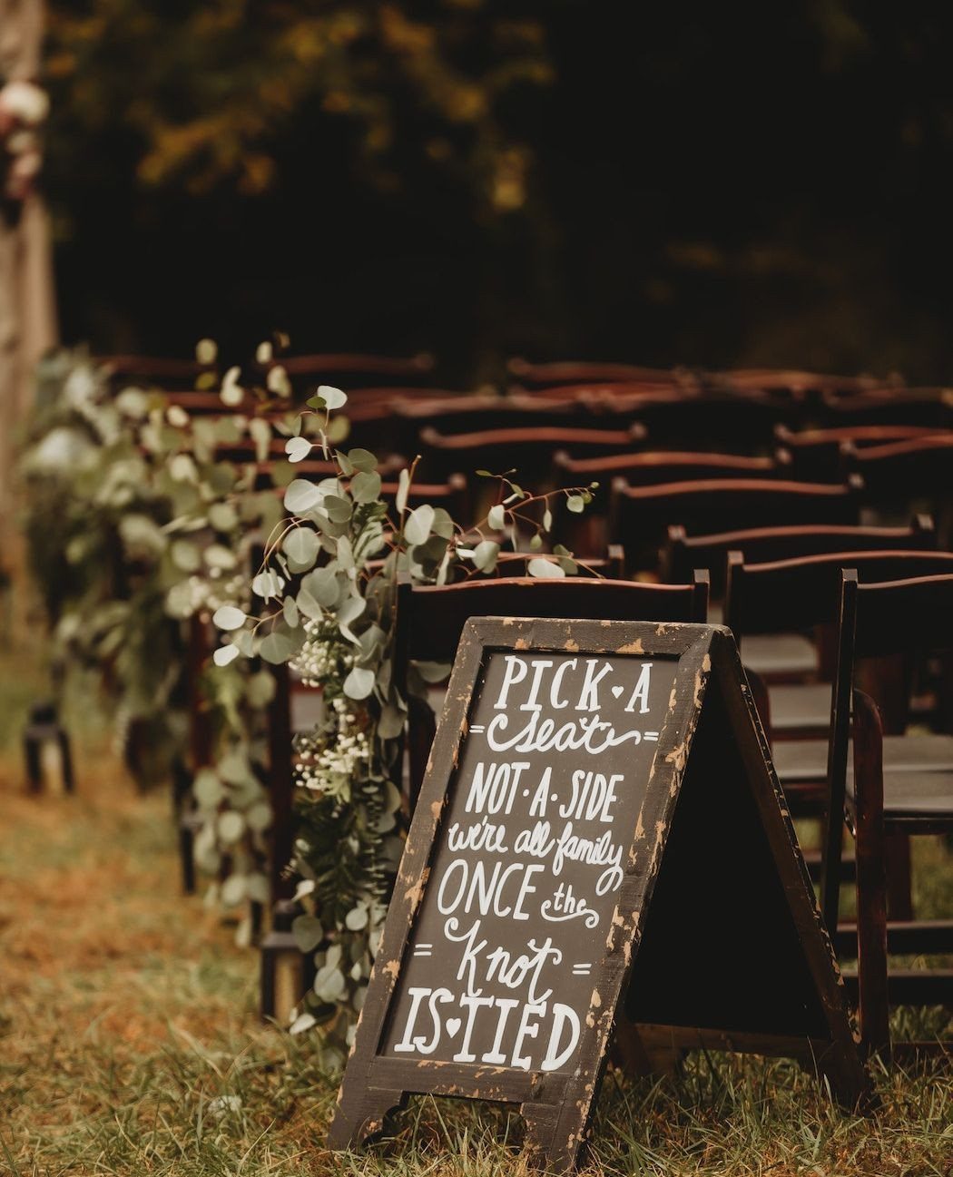 A wooden chalkboard sign with funny wedding seating text sits before rows of outdoor chairs.