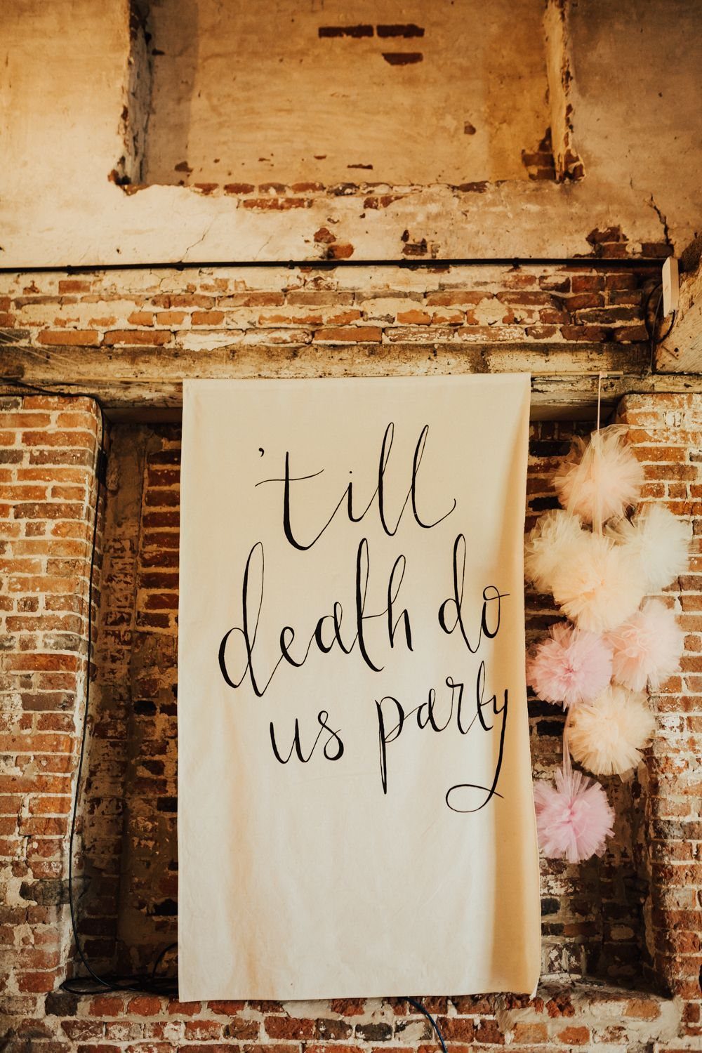 A cream fabric banner with black calligraphy reading till death do us party hangs on brick.