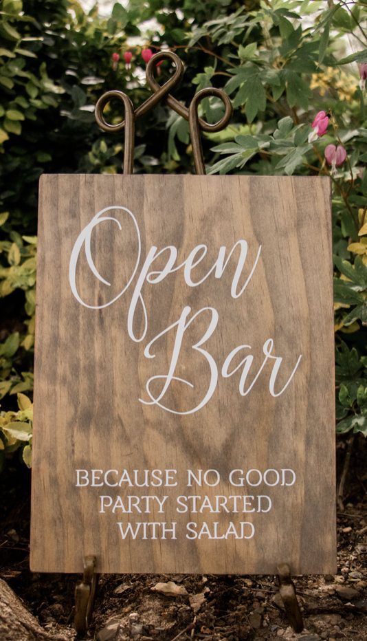 A wooden sign with white text reading Open Bar because no good party started with salad.