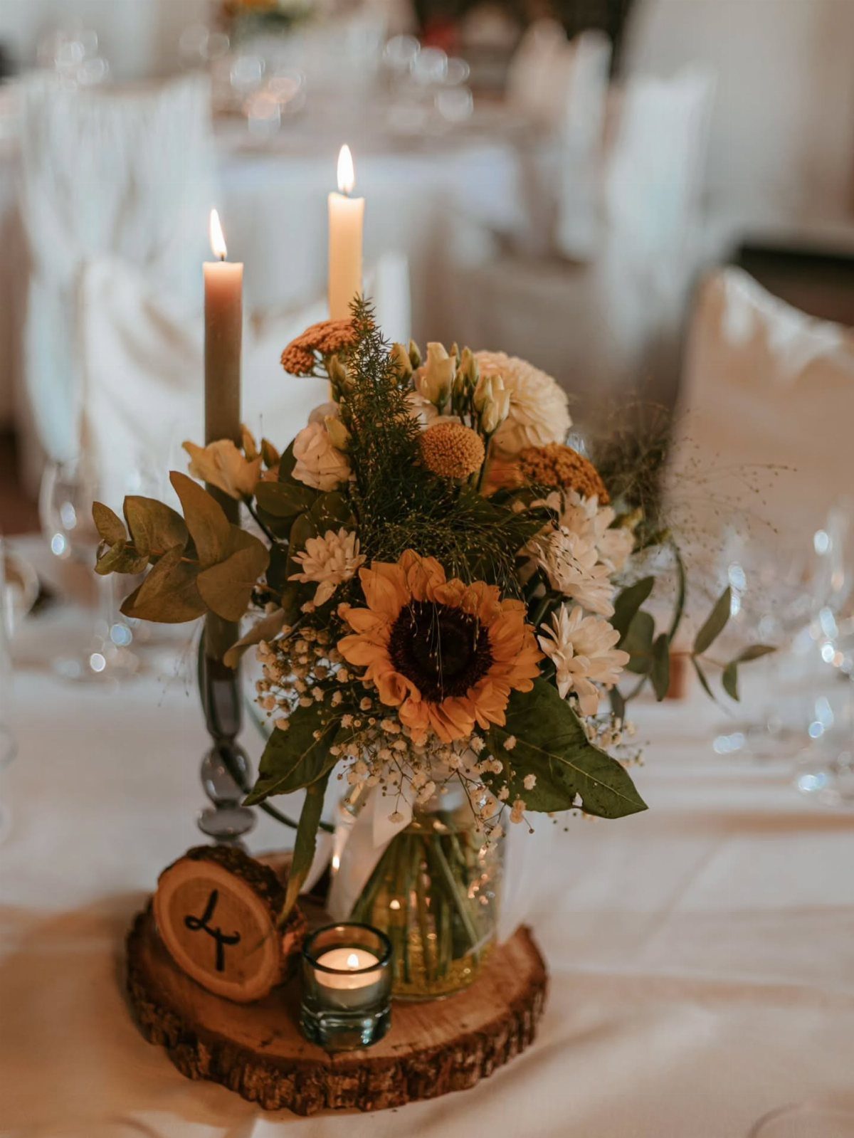 Sunflower centerpiece with white flowers and greenery on a wooden slice with a candle.
