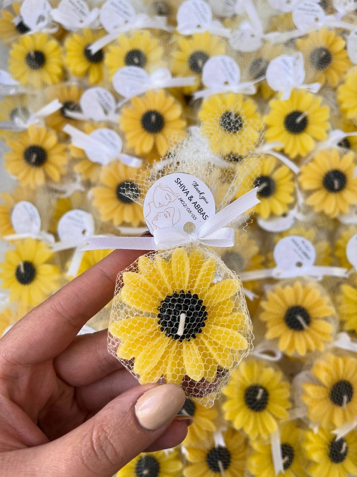 A hand holding a sunflower-shaped candle wrapped in white netting with a personalized tag.