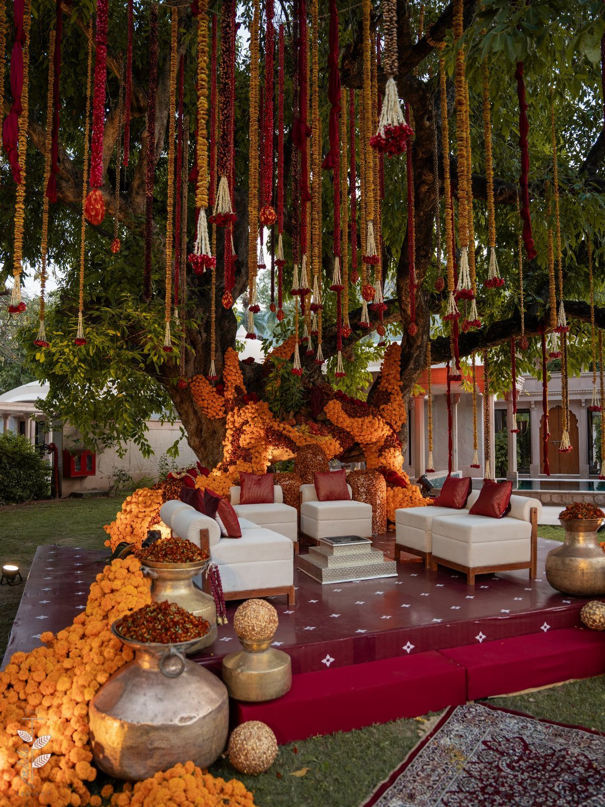 7 Wedding Tree Decorations That Work Outside the Ballroom 6 Flower garlands hang from a large tree over a decorated outdoor lounge with white sofas.