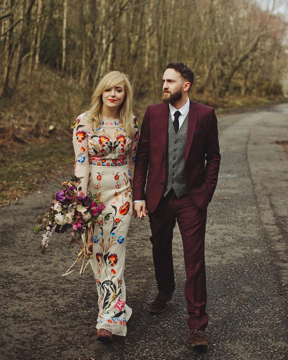 A woman in a floral embroidered dress and a man in a maroon suit walking outdoors.