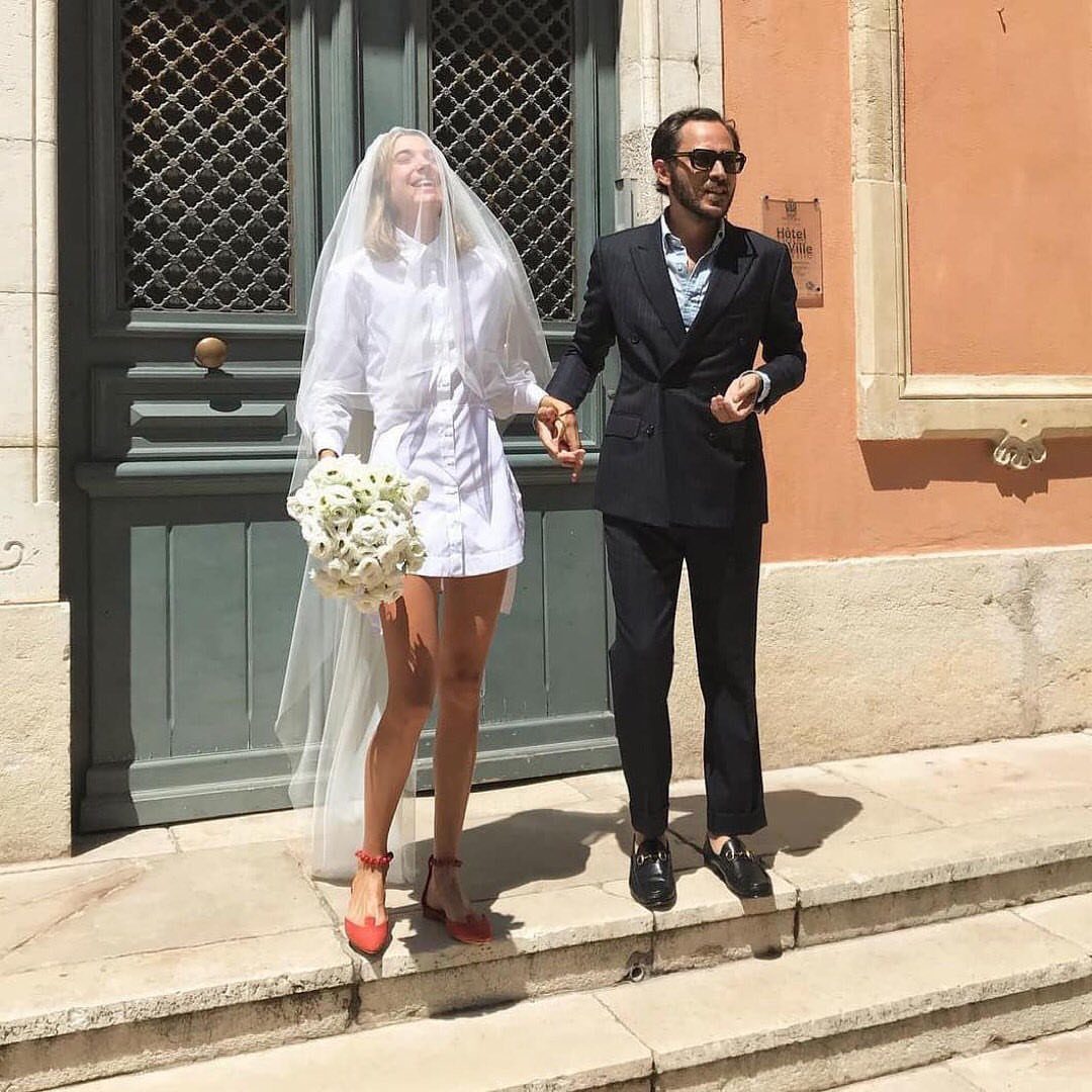 A bride in a short white shirt dress and long veil walking with a groom.
