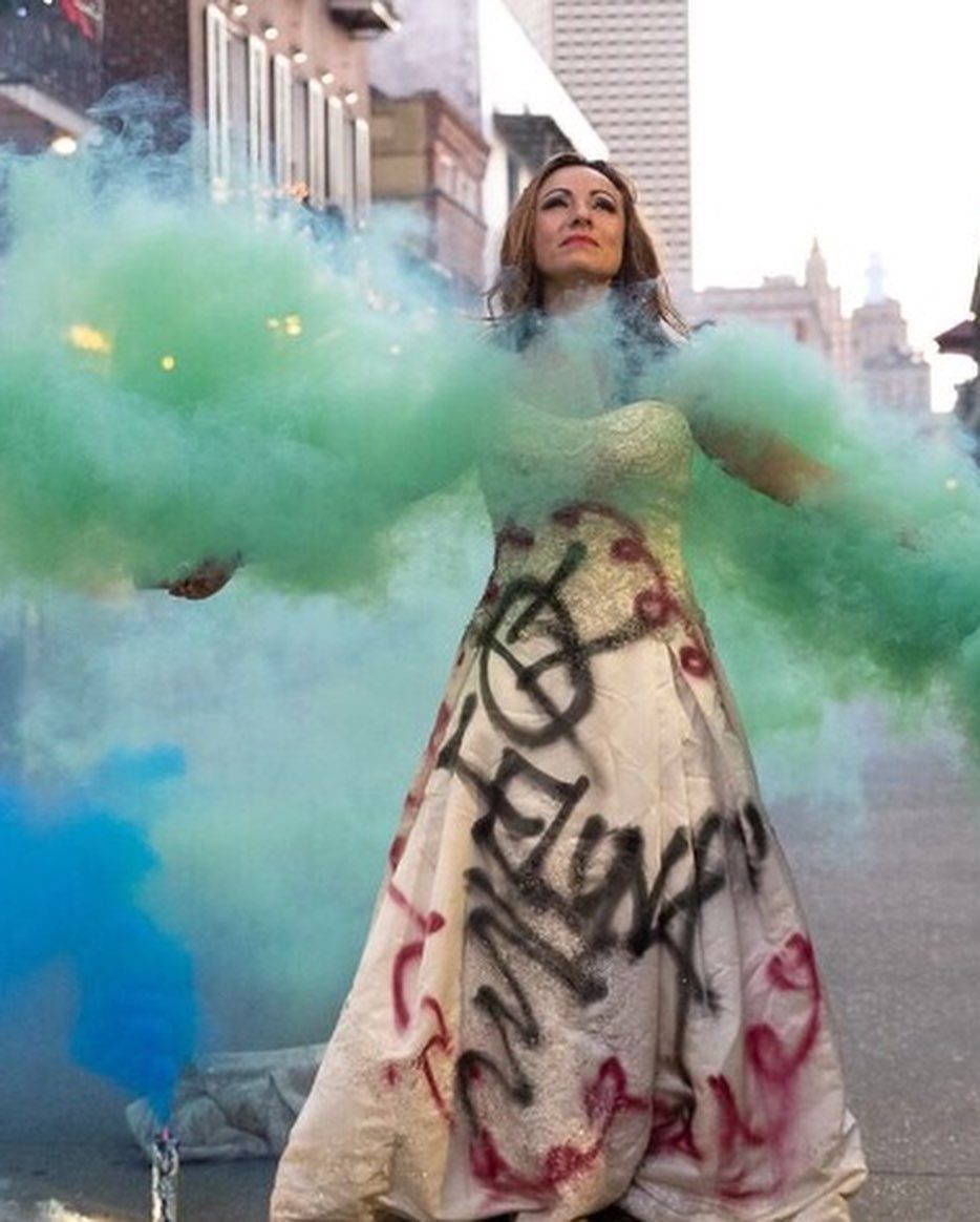 Woman in a spray-painted white wedding dress standing amidst clouds of teal and blue smoke.