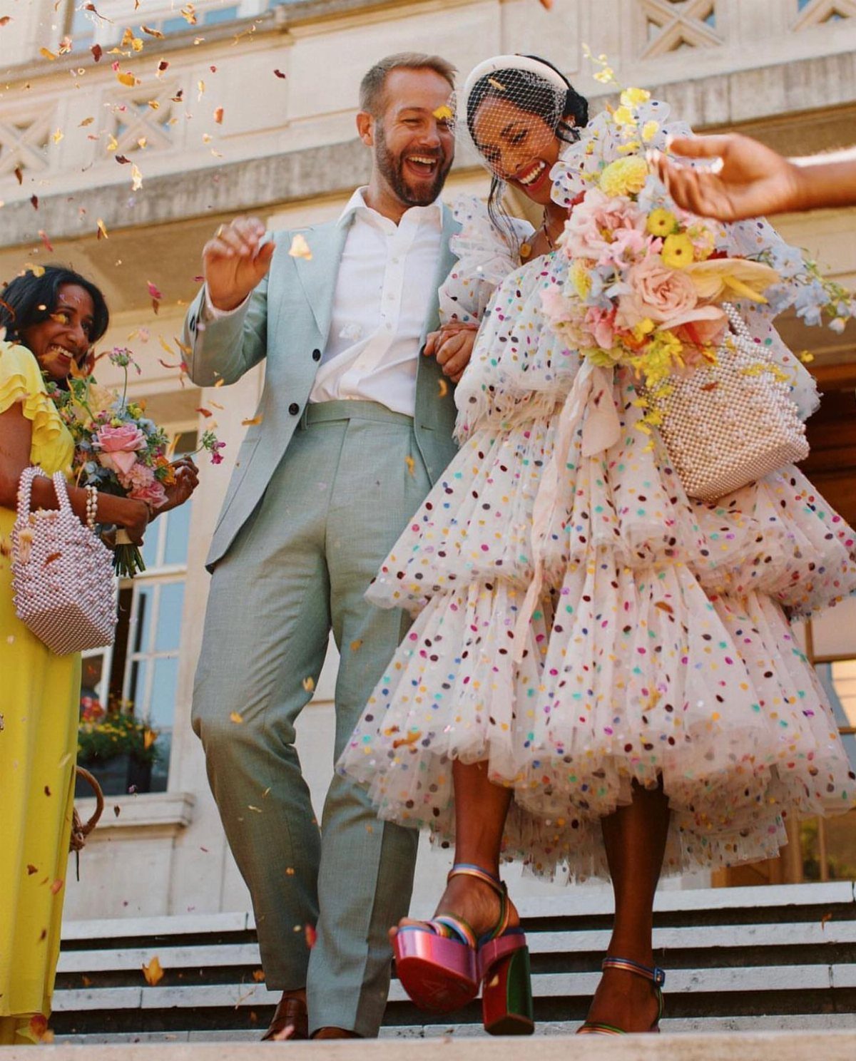A laughing bride in a colorful confetti dress and a groom in a light blue suit celebrate outdoors.