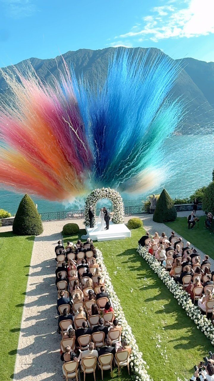 A wedding ceremony with a floral arch and massive colorful plumes overlooking a lake and mountains.