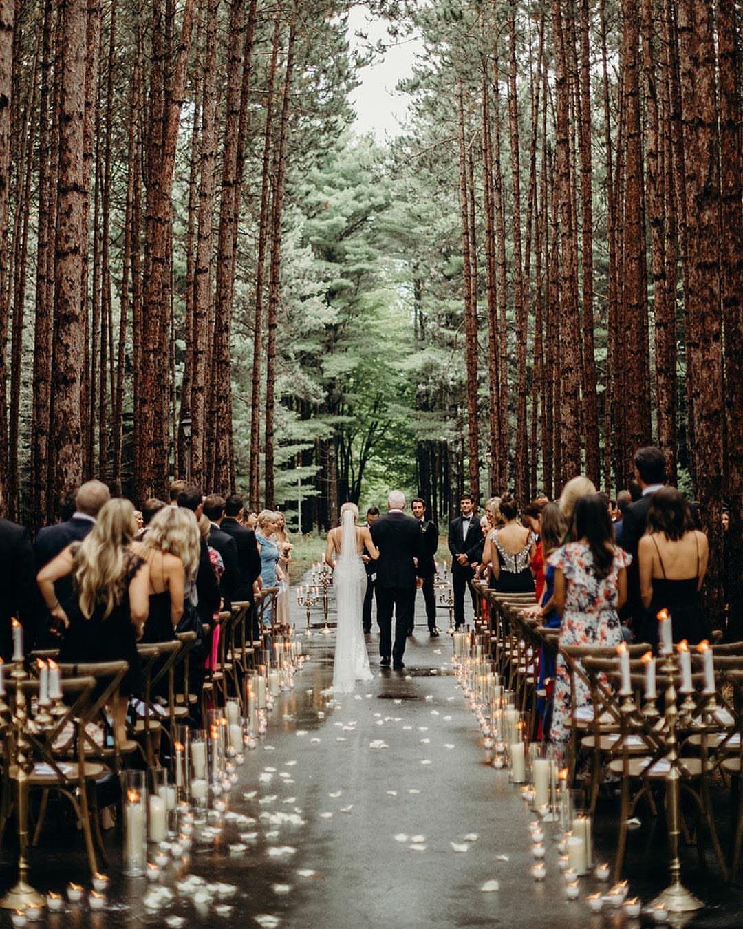 A bride and groom walk down a candlelit forest aisle during a wedding ceremony.