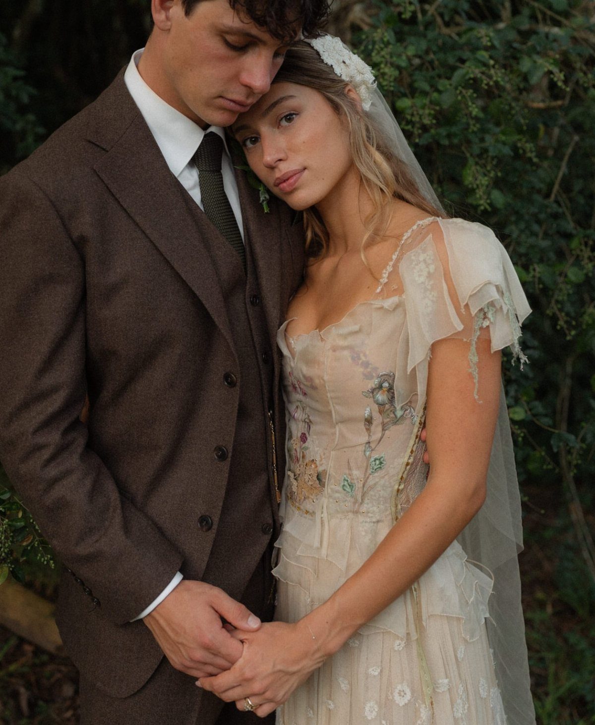 A couple posing together, the woman wearing a cream dress with floral appliques.