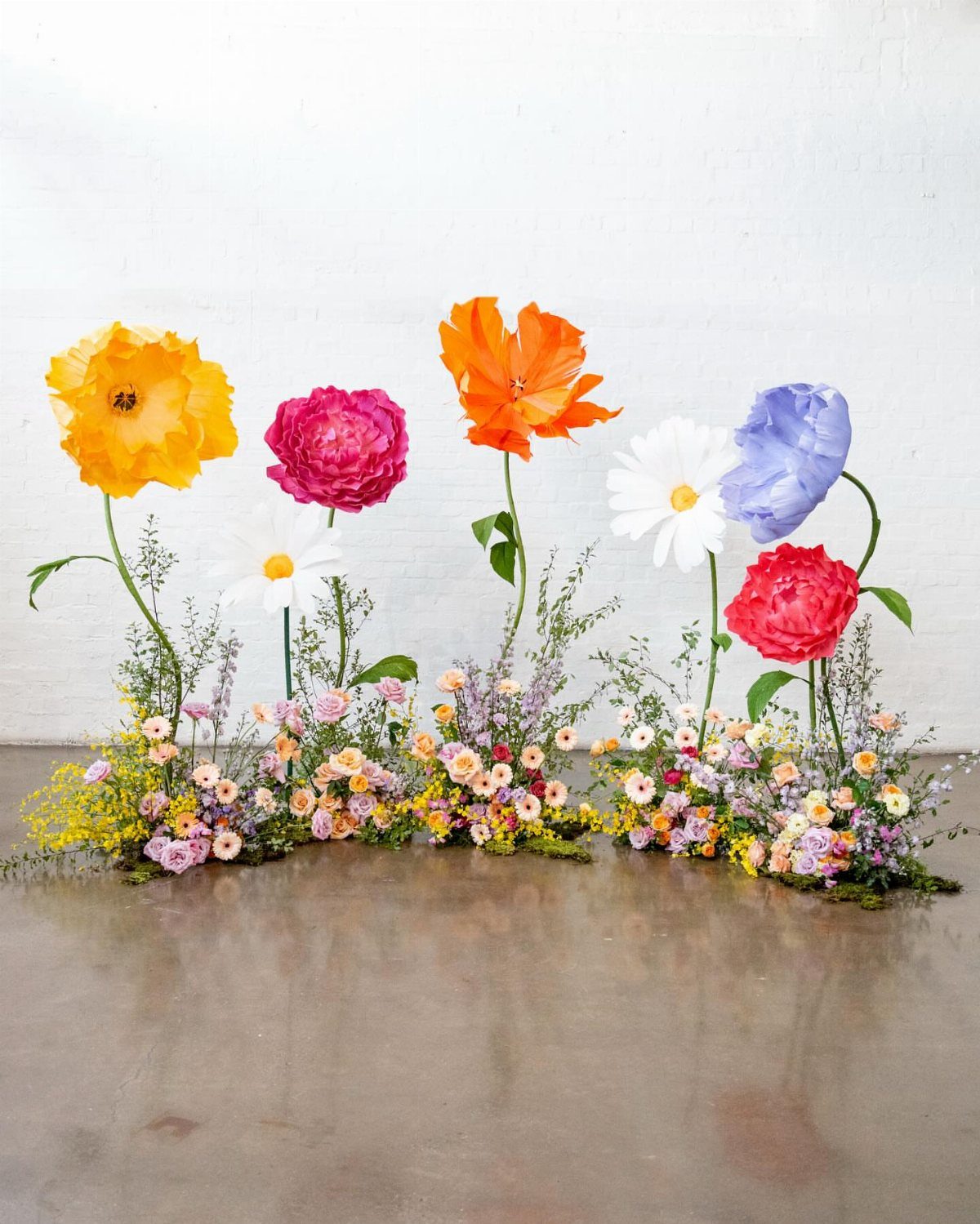 Oversized colorful artificial flowers stand above a lush arrangement of small wildflowers against a white wall.
