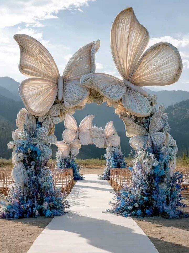 Large white butterfly sculptures arching over a white wedding aisle lined with blue flowers.