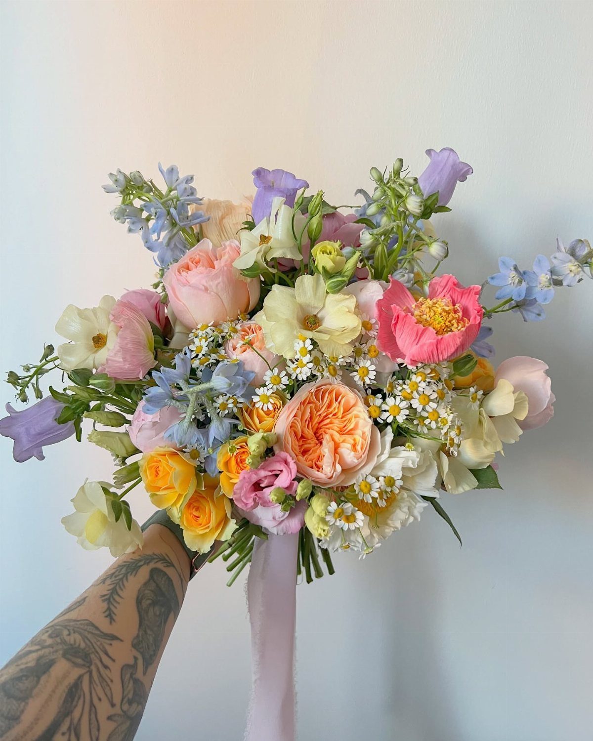 A colorful floral bouquet with large pink roses, yellow flowers, and blue accents held against a white wall.