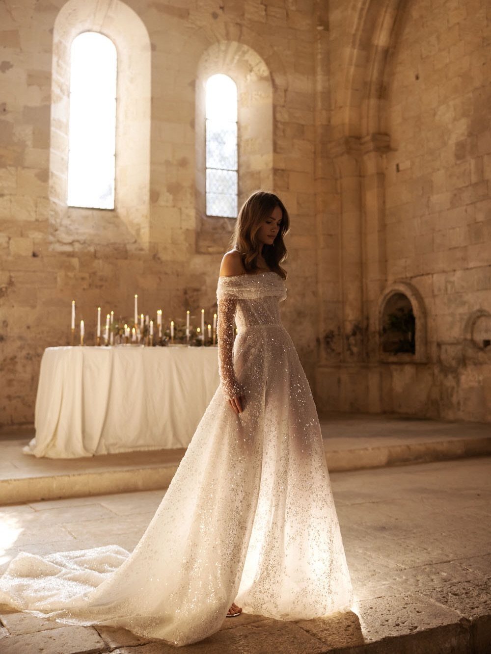 19 Winter Wonderland Wedding Ideas for a Modern Celebration 18 A bride in a shimmering white gown stands in a sunlit stone cathedral with candles.