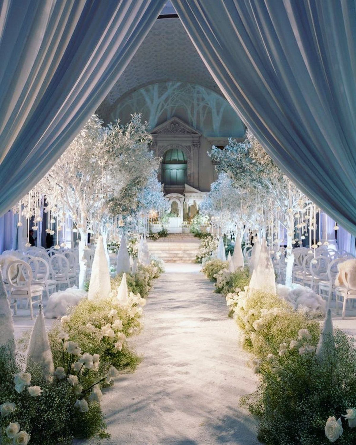 19 Winter Wonderland Wedding Ideas for a Modern Celebration 2 A winter wedding aisle lined with white flowering trees and baby's breath under blue drapes.